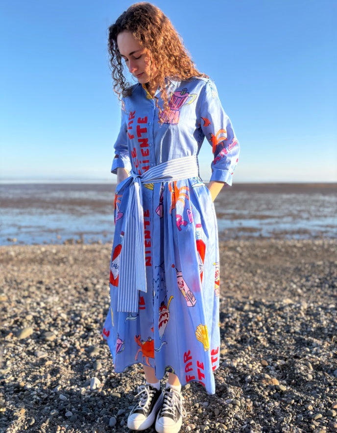 Woman standing on a beach wearing a colourful dress with a pattern of coffee cups and saucers.