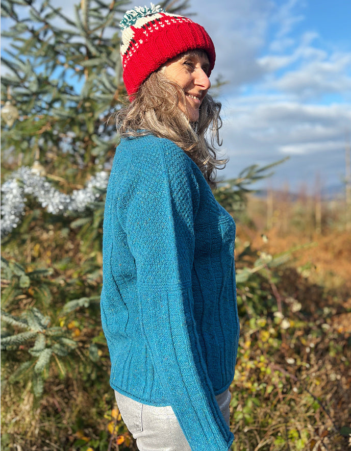 Person wearing a teal blue cable sweater and red beanie outdoors with trees and sky in the background
