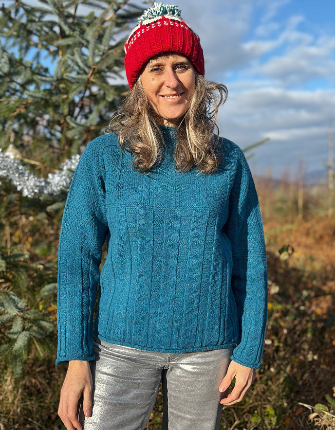 Person wearing a teal blue cable sweater and red beanie outdoors with trees and sky in the background