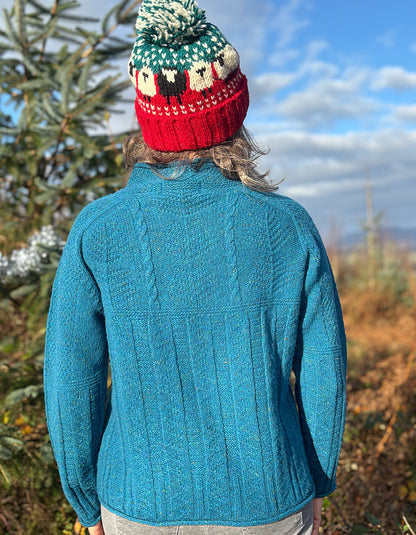 Person wearing a teal blue cable sweater and red beanie outdoors with trees and sky in the background