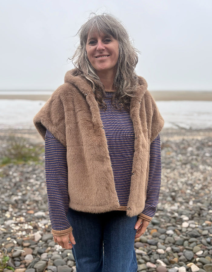 A woman with gray hair stands on a pebble beach wearing a tan faux fur hooded vest over a blue and red striped long-sleeve shirt and blue jeans.