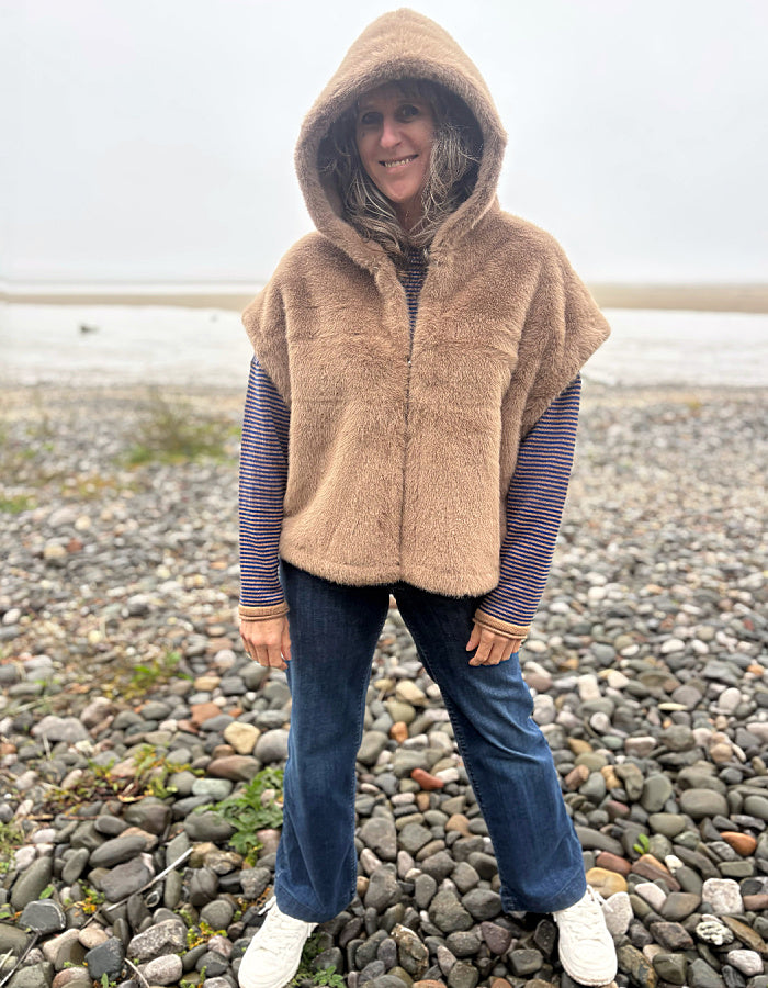 A woman wearing a tan fuzzy hooded poncho vest over a striped long-sleeve shirt, blue jeans, and white sneakers, standing on a rocky beach.