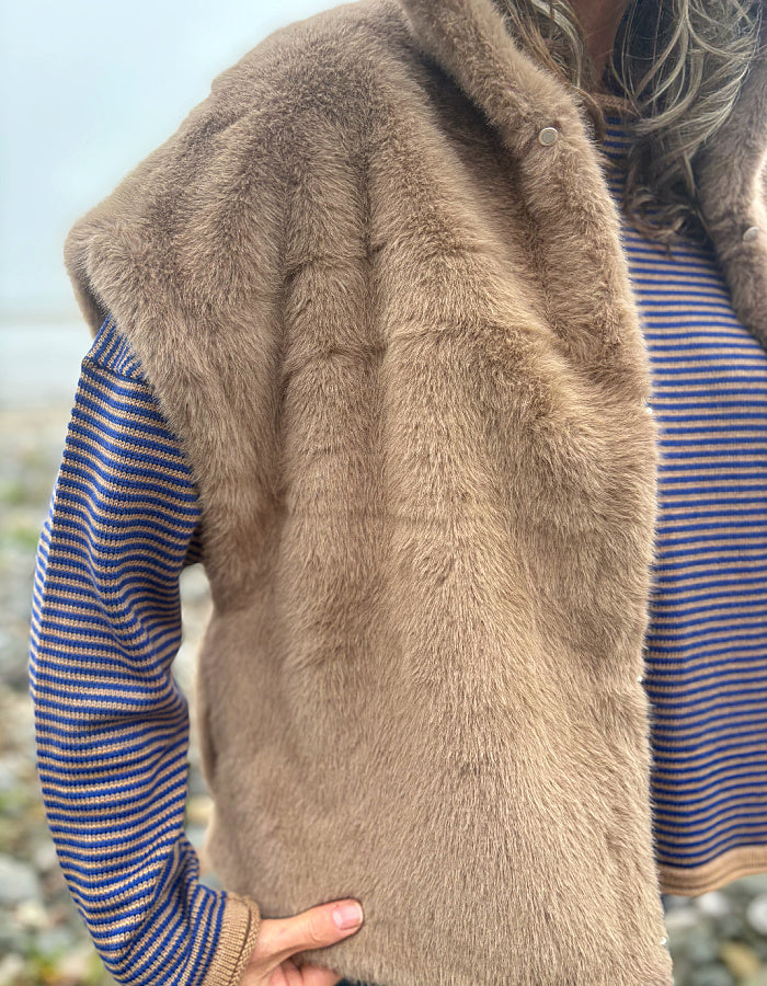 A close-up of a person wearing a brown faux fur vest over a blue and orange striped knit sweater, with a blurred outdoor background.