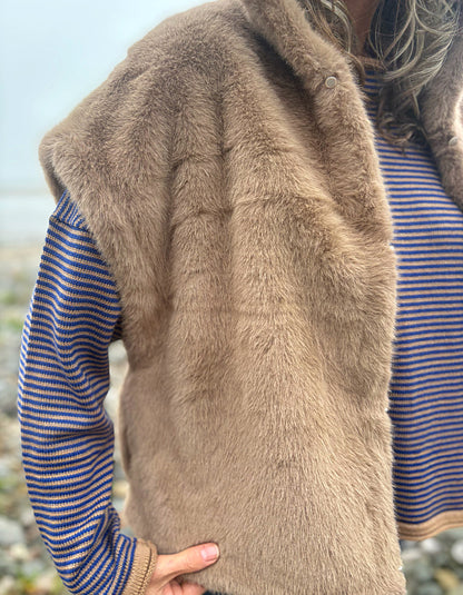 A close-up of a person wearing a brown faux fur vest over a blue and orange striped knit sweater, with a blurred outdoor background.