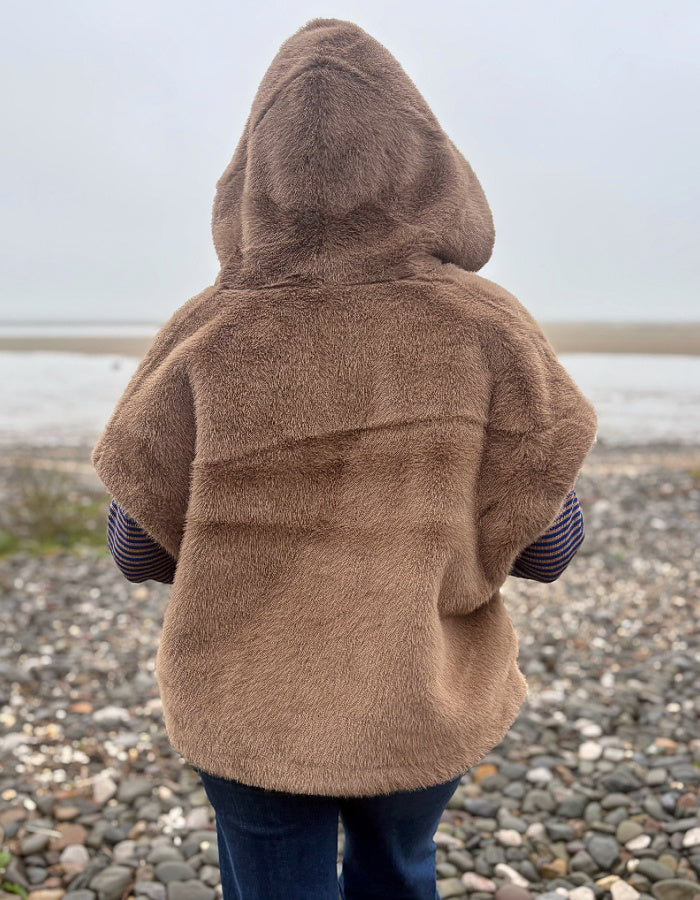 A person viewed from behind wearing a brown fuzzy hooded jacket on a pebble beach, with water and a pale sky in the background.