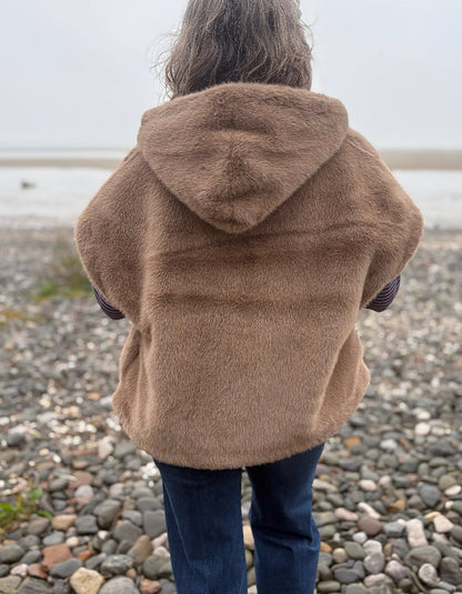 A person viewed from behind wearing a brown fuzzy hooded jacket and blue jeans, standing on a pebble beach with water in the background.