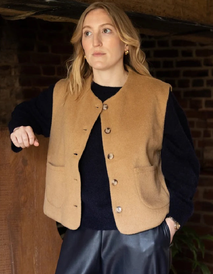 A woman wearing a tan wool-blend vest over a black sweater and dark leather pants, standing in a rustic indoor setting with a brick and wood wall background.
