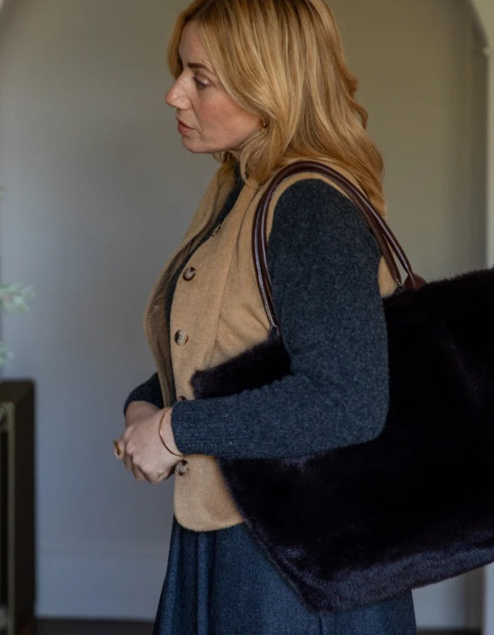 A woman in a beige button-front vest over a dark blue sweater and skirt carries a large black faux-fur shoulder bag with dark straps in an indoor setting.
