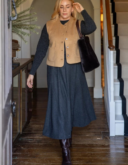 A woman walking down a wooden staircase hallway wearing a tan suede vest over a dark gray long-sleeve top and a dark gray midi skirt, carrying a black fur-trimmed shoulder bag.