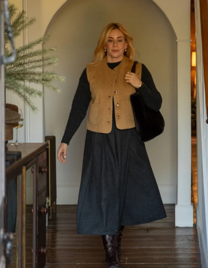 A woman walks through an indoor hallway wearing a tan suede vest over a black long-sleeve top and a dark charcoal midi skirt, with dark brown boots and a black shoulder bag.