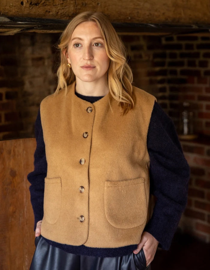 A woman wearing a tan wool-blend button-front vest over a dark navy sweater, standing in a rustic room with a brick wall and wooden ceiling.