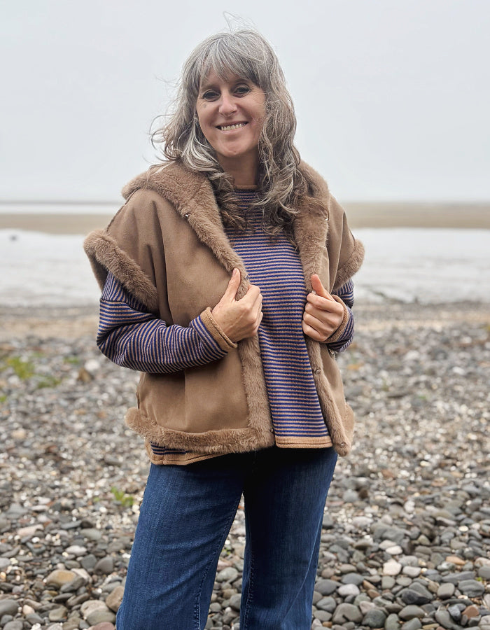 A woman with gray hair stands on a pebble beach wearing a tan shearling-style vest with brown faux-fur trim over a purple and blue striped long-sleeve top and blue jeans.