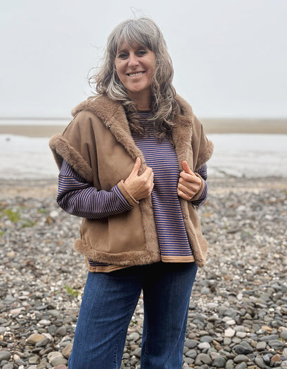 A woman with gray hair stands on a pebble beach wearing a tan shearling-style vest with brown faux-fur trim over a purple and blue striped long-sleeve top and blue jeans.