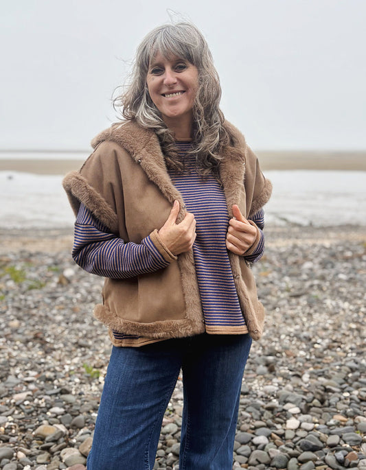 A woman with gray hair stands on a pebble beach wearing a tan shearling-style vest with brown faux-fur trim over a purple and blue striped long-sleeve top and blue jeans.