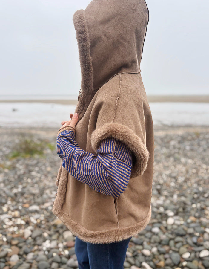 A person standing on a pebble beach wearing a brown suede-like hooded vest with fuzzy trim over a blue and red striped long-sleeve shirt and blue jeans.