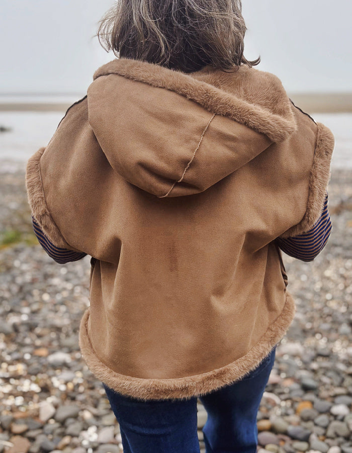 A person viewed from behind wearing a tan hooded jacket with a soft brown faux-fur trim, standing on a pebble beach.