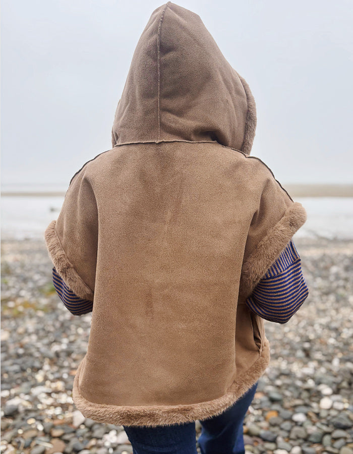 A child viewed from behind wearing a tan suede-like hooded vest with fuzzy trim over a striped shirt, standing on a pebble beach.