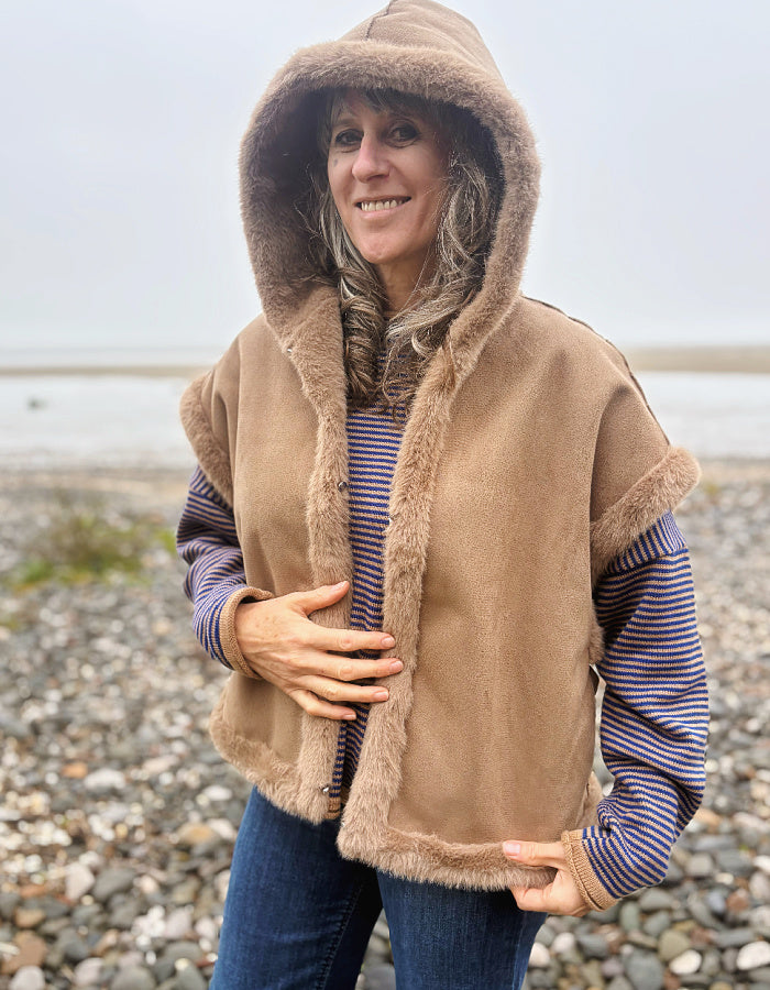 A woman wearing a tan hooded shearling-style vest over a blue and white striped shirt and blue jeans, standing on a pebble beach with water in the background.
