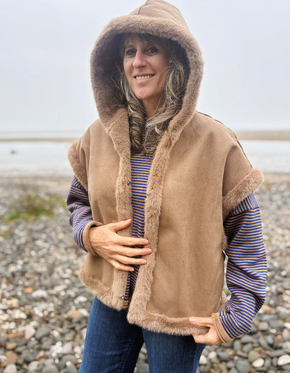 A woman wearing a tan hooded shearling-style vest over a blue and white striped shirt and blue jeans, standing on a pebble beach with water in the background.