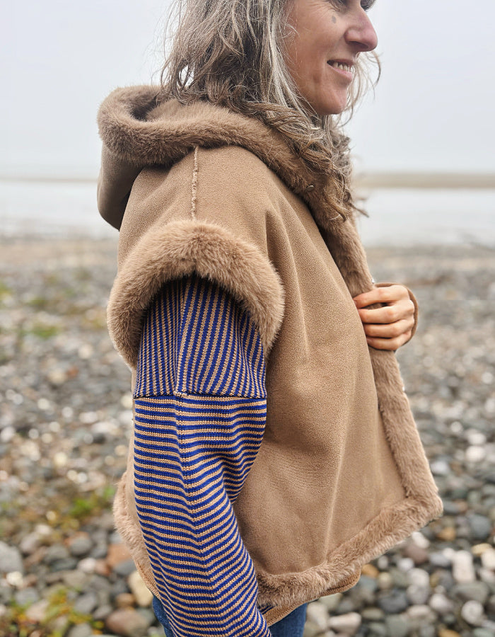 A woman wearing a tan fleece hooded vest with brown faux-fur trim, layered over a blue and red striped knit sweater, standing on a pebble beach.