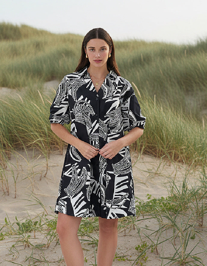 Woman wearing a black and white patterned dress standing on a sandy beach with grasses.