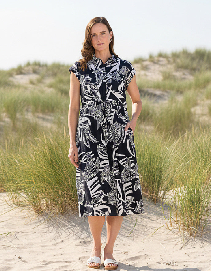 Woman wearing a black and white patterned dress on a sandy beach with grass.