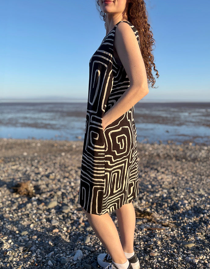 Woman in a black and white patterned dress standing on a pebbly beach with ocean in the background.