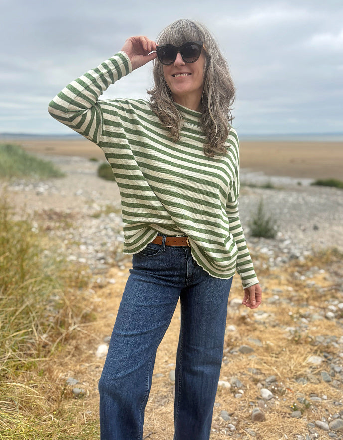 A woman wearing a green and white striped long-sleeve knit top and blue jeans stands outdoors on a rocky beach or shoreline under an overcast sky.
