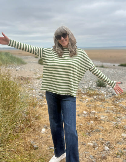 A woman with gray hair and sunglasses stands on a rocky beach with her arms outstretched, wearing a green and white striped long-sleeve knit sweater, blue jeans, and white sneakers.