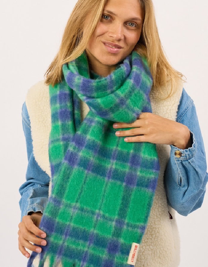 A woman wearing a green and blue plaid scarf wrapped around her neck, styled with a blue denim jacket and cream sherpa vest against a plain white background.