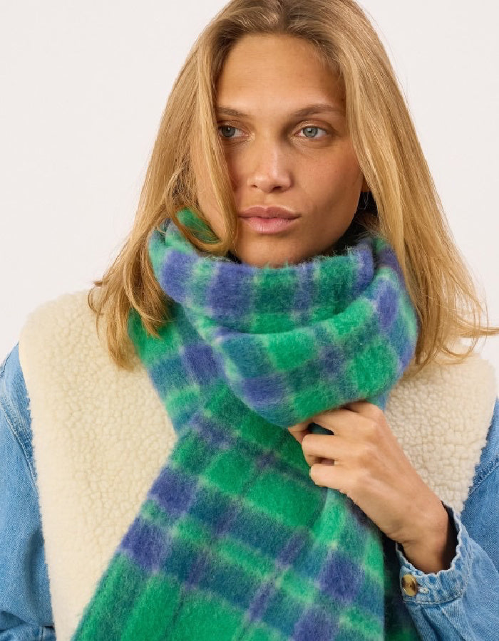 A woman wearing a plush green and blue plaid scarf wrapped around her neck, paired with a cream sherpa vest and a light blue denim jacket against a plain white background.