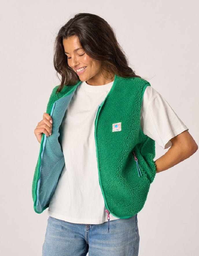 A woman wearing a green sherpa fleece vest over a white t-shirt and blue jeans, holding the vest open to show the teal interior lining. The vest features a small square logo patch on the left chest.