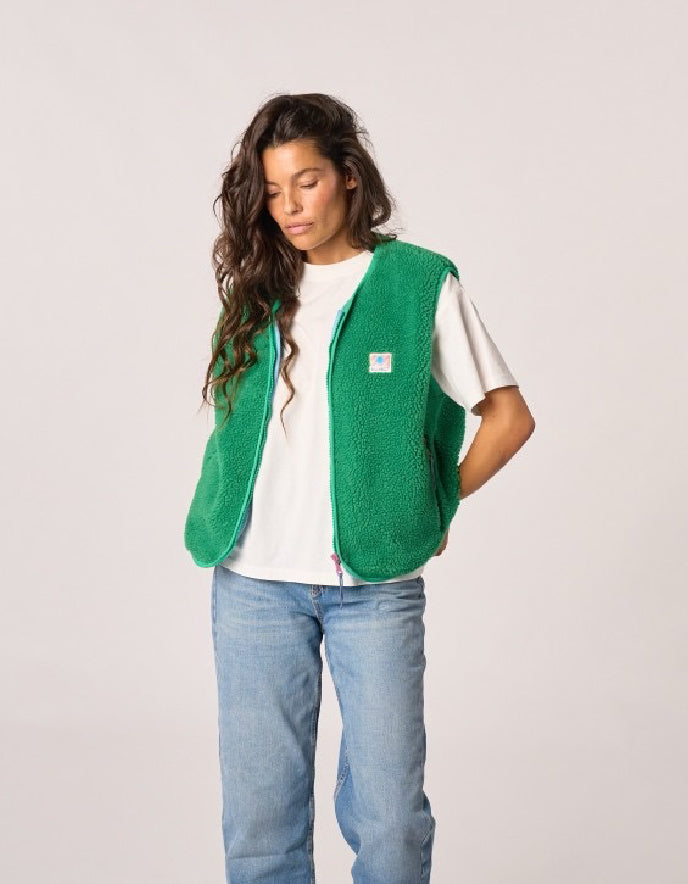 A woman wearing a bright green fuzzy fleece vest over a white t-shirt and light blue jeans, standing against a light gray studio background.