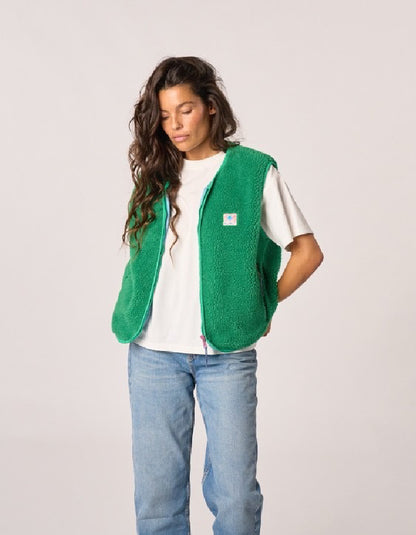A woman wearing a bright green fuzzy fleece vest over a white t-shirt and light blue jeans, standing against a light gray studio background.