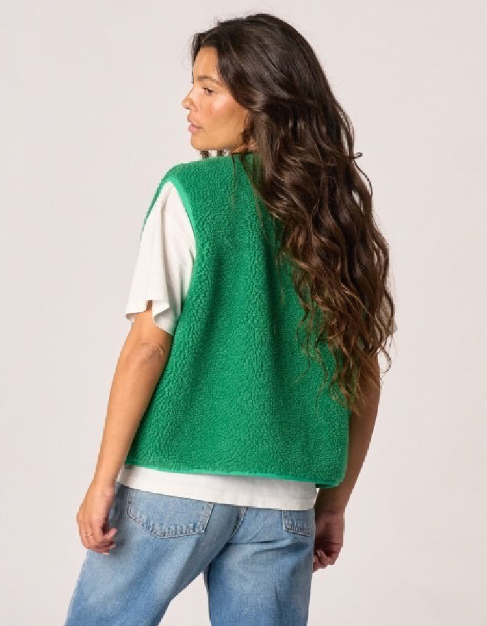 A woman shown from the back wearing a bright green textured sleeveless vest over a white t-shirt and blue jeans, against a light gray studio background.