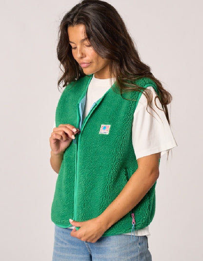 A woman wearing a bright green fleece zip-up vest over a white t-shirt and blue jeans, shown against a plain light gray studio background. The vest features a small embroidered Carhartt logo on the left chest.