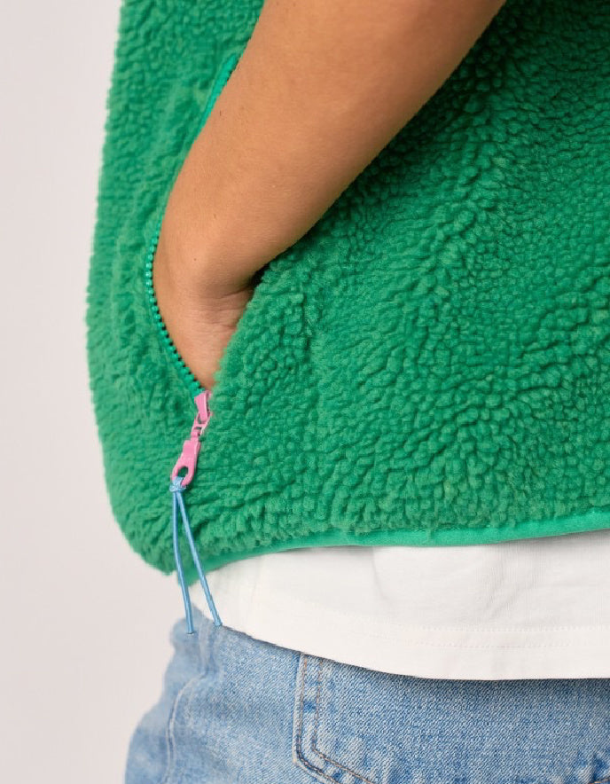 Close-up of a person wearing a bright green sherpa fleece zip-up vest over a white shirt and light blue jeans, with the hand in the pocket and a pink zipper pull visible.