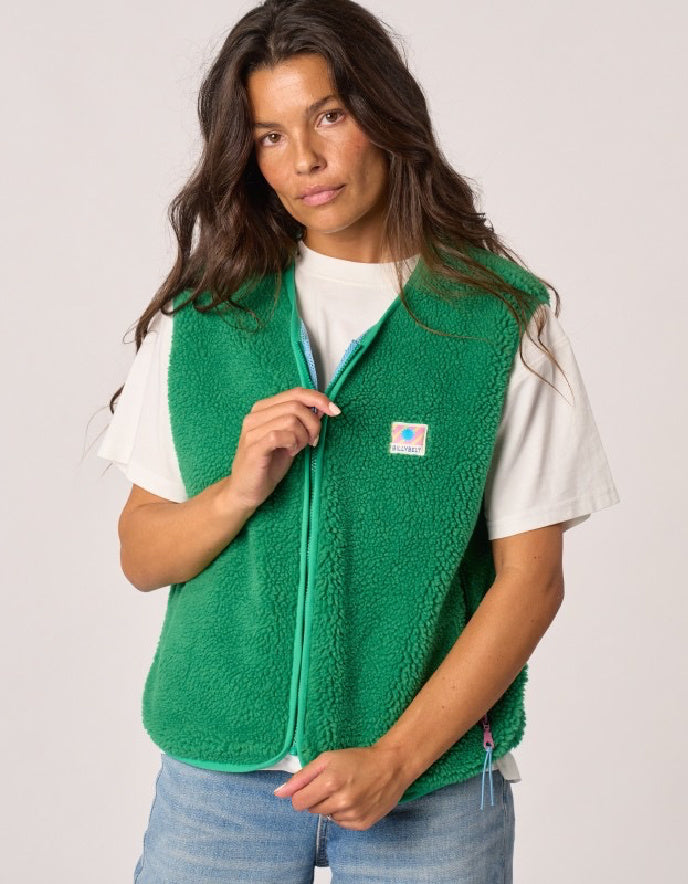 A woman wearing a bright green fleece zip-up vest over a white t-shirt and blue jeans, photographed against a light gray studio background. The vest features a small square logo patch on the left chest.