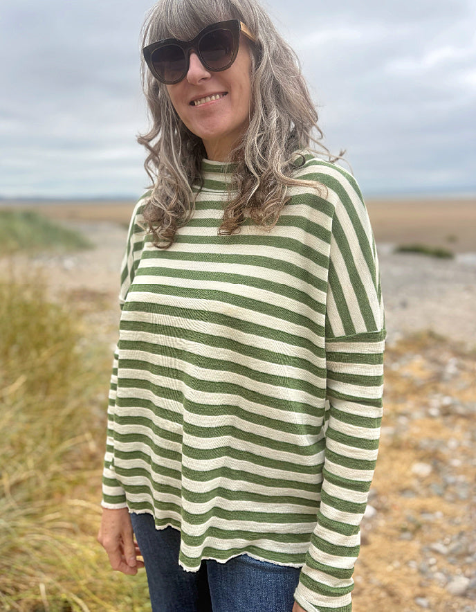 A woman wearing a green and cream striped long-sleeve knit top and blue jeans, standing outdoors on a beach or dune path under an overcast sky.