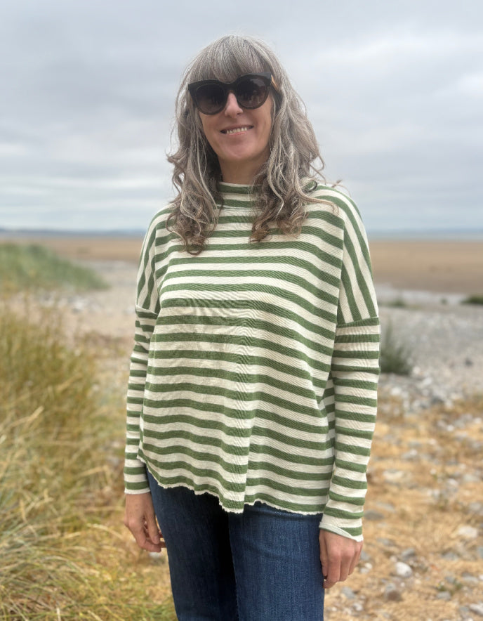 A woman with gray hair and sunglasses wearing a green and cream striped long-sleeve top, standing outdoors on a beach path.