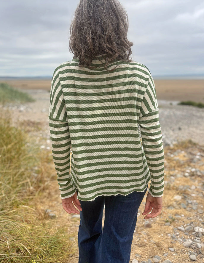 A person standing outdoors on a rocky beach or dune path, viewed from behind, wearing a green and cream striped long-sleeve top and dark blue jeans under an overcast sky.