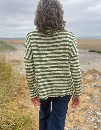 A person standing outdoors on a rocky beach or dune path, viewed from behind, wearing a green and cream striped long-sleeve top and dark blue jeans under an overcast sky.