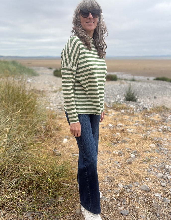 A woman wearing a green and white striped long-sleeve top, dark blue flared jeans, and white sneakers stands on a rocky, grassy beach under an overcast sky.