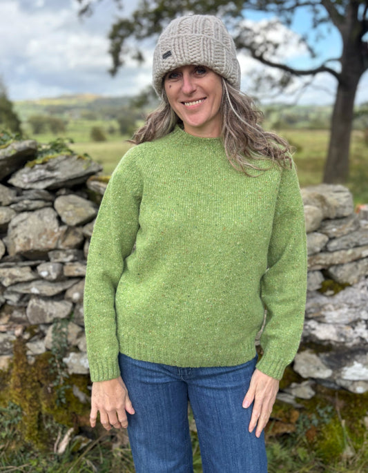 Woman wearing a green sweater and beige hat standing in a natural setting with stone walls and trees.