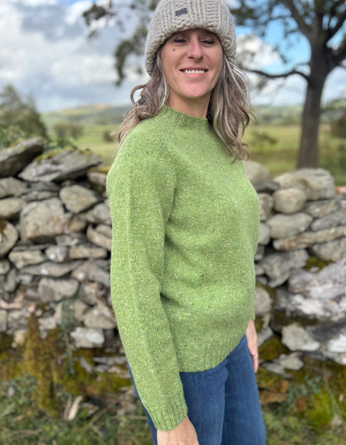Woman wearing a green sweater and beige beanie standing in front of a stone wall with a natural landscape background.