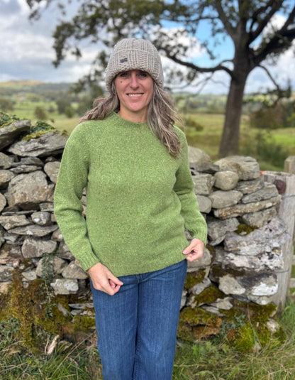Person wearing a green sweater and gray beanie standing in front of a stone wall with a scenic background.