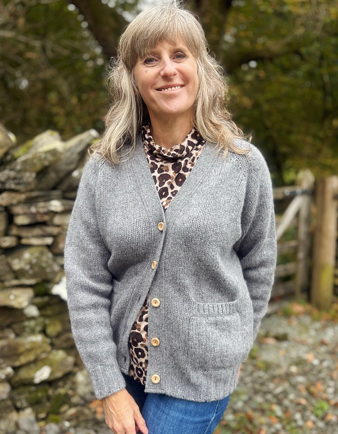 Woman wearing a gray cardigan over a leopard print top, standing outdoors with a stone wall and trees in the background.