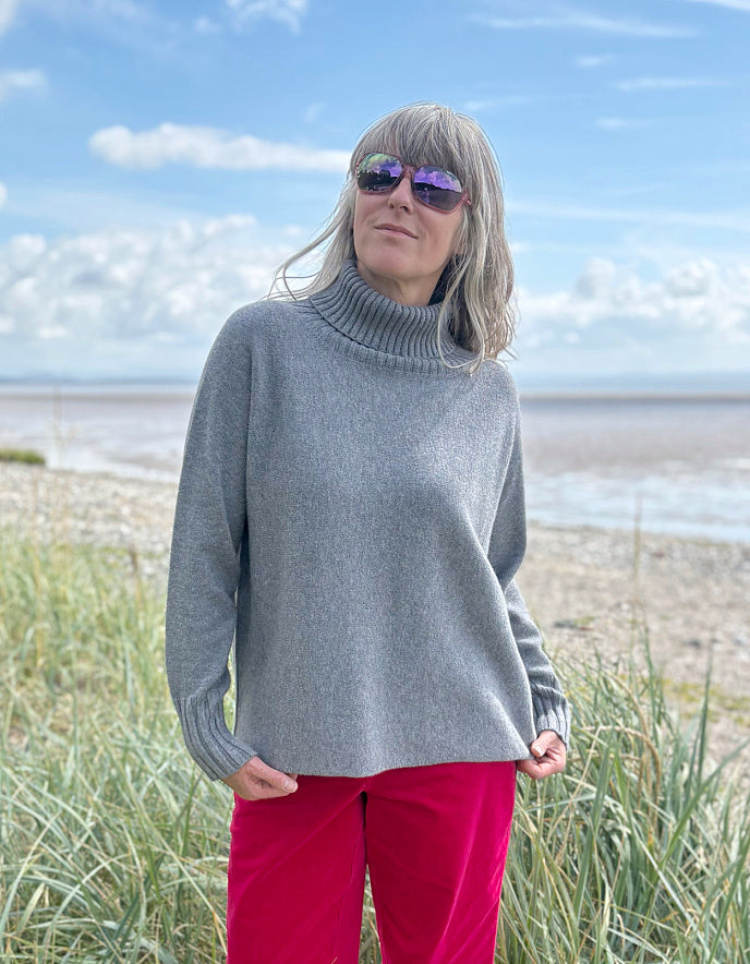 Person wearing a grey turtle neck sweater and sunglasses standing in a grassy field with a blue sky.