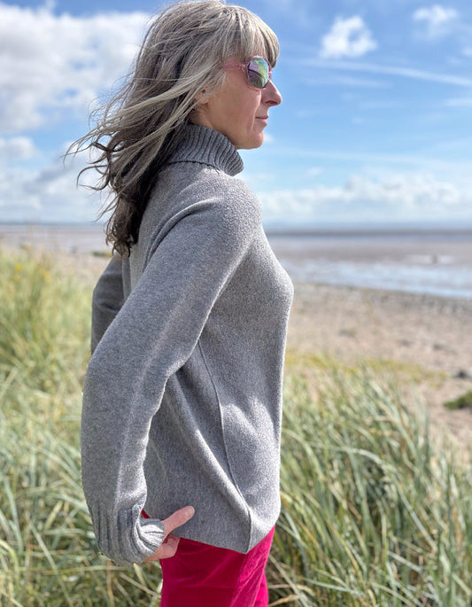 Person wearing a grey turtle neck sweater and sunglasses standing in a grassy field with a blue sky.
