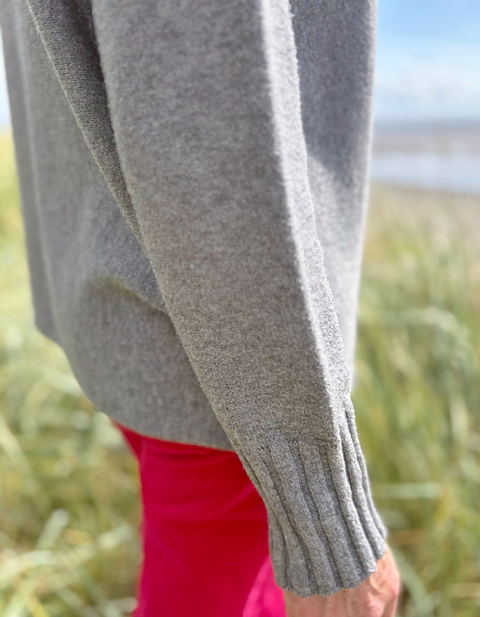 Person wearing a grey turtle neck sweater and sunglasses standing in a grassy field with a blue sky.